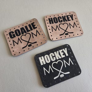 May include: Three square faux leather patches with pre-punched holes along the edges. Two patches are light tan with black text reading GOALIE MOM and HOCKEY MOM. One patch is black with white text reading HOCKEY MOM, each featuring a heart-shaped hockey stick.