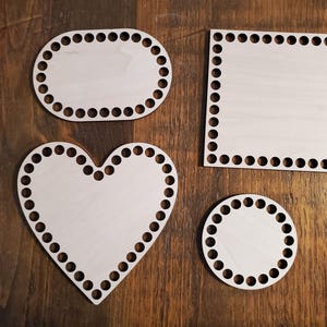 May include: Four wooden loom knitting boards in different shapes: oval, square, heart, and circle. Each board has a white surface and a black border of evenly spaced holes.