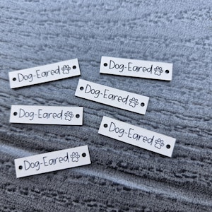 May include: Five rectangular silver-toned metal tags, each featuring the text "Dog-Eared" in a black sans-serif font followed by a small black paw print icon. Each tag has a small circular hole punched at both the left and right ends for attachment.