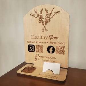 May include: A light-coloured wooden display sign featuring the text Healthy Glow, Natural, Vegan, Sustainable, PETA APPROVED, and social media icons with QR codes. The base includes a square tap-to-pay area and a slot holding white business cards.