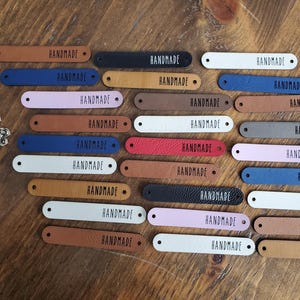 May include: A collection of twenty-four leather labels with the word "HANDMADE" printed in black on each label. The labels are in various colours, including brown, white, pink, red, blue, and grey.
