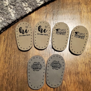 May include: Six oval-shaped beige leather soles with pre-punched holes along the edges. Two soles feature the text 'hi I'm new here' with a heart, two feature 'little peanut' with a heart, and two feature 'DADDY'S HOCKEY BUDDY' with graphic accents.