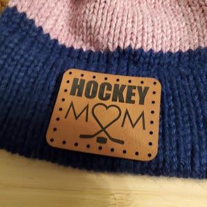 May include: A blue and pink knitted beanie with a brown leather patch that says "HOCKEY MOM" with a heart and hockey sticks.