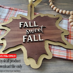 May include: A wooden leaf-shaped sign with a gold outline. The sign says "FALL sweet FALL" in white letters. The sign is on a plaid fabric and a wooden surface.