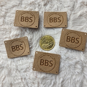 May include: Five tan rectangular labels with the embossed text "BIG BUTTON SCARF" and "BBS" inside a circle. A gold Canadian dollar coin is in the center. The labels are on a white, fluffy surface.