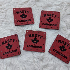 May include: Five square red leather labels with pre-punched holes along the edges. Each label features the black text NASTY CANADIAN and a black maple leaf graphic with a small heart cutout in the centre of the leaf, printed on the surface.