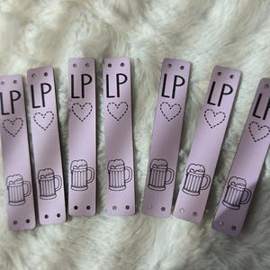 May include: Seven rectangular light purple tags, each featuring the letters LP at the top, a dashed-line heart icon in the centre, and a black line drawing of a beer mug at the bottom. Each tag has two small circular holes at both ends.