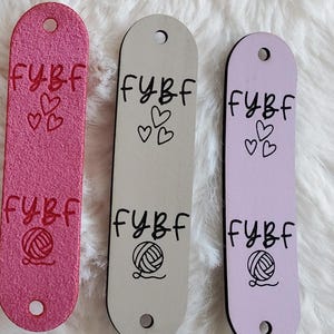 May include: Three elongated rectangular tags in pink, beige, and light purple. Each tag features two circular holes at the ends and is imprinted with the text FYBF, a cluster of three small hearts, and a graphic illustration of a ball of yarn.