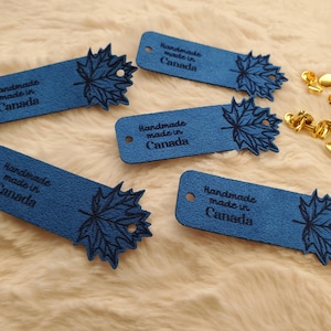 May include: Five blue leather tags with a maple leaf design and the text "Handmade made in Canada". The tags are rectangular with a hole at one end. Gold-colored rivets are also visible.