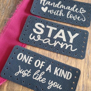 May include: Three black leather labels with gold lettering. The first label says "Handmade with love" with a heart symbol. The second label says "Stay warm". The third label says "One of a kind just like you".