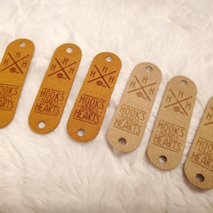 May include: Six elongated oval tags in two shades of tan, each featuring a laser-engraved design with the text "HOOKS HANDS HEARTS" and a crossed hook symbol with the letter H in each quadrant. Each tag has a circular hole at the top and bottom.
