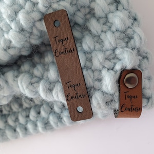 May include: A light blue chunky knit hat featuring two brown leather labels. One long rectangular label has two holes and the text Toque Couture, while a smaller folded label is secured with a silver metal rivet and also displays the text Toque Couture.