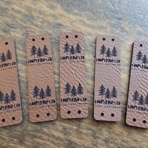 May include: Five rectangular tan faux leather clothing labels with rounded corners. Each label features two pre-punched holes at the top and bottom, with a black printed design of three pine trees and the text "LOOPY KNITS CO COZY KNITWEAR".