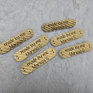 Peut inclure: Cinq étiquettes rectangulaires beige clair avec un motif texturé semblable à un tissage. Chaque étiquette comporte deux petits trous aux extrémités et le texte imprimé en noir 'made by me Marissa G' centré, disposées en éparpillement.