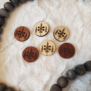 Peut inclure: Six boutons en bois ronds avec un motif de forêt. Les boutons présentent un motif d'arbre et de trous découpés au laser. Certains sont de couleur claire, d'autres sont brun foncé. Les boutons sont disposés sur une surface blanche et duveteuse.