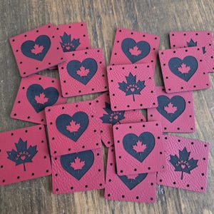 May include: A collection of square red faux leather tags featuring a black heart shape with a small red maple leaf inside. Some tags display a black maple leaf with the word CANADA and a tiny heart printed on the surface. Each tag has pre-punched holes.
