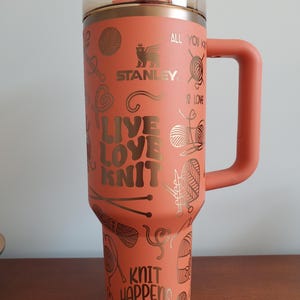 Peut inclure: Un gobelet en acier inoxydable de couleur corail avec une poignée et une paille. Le gobelet présente un motif doré avec les mots "Live Love Knit" et "Knit Happens" ainsi que des images de laine, d'aiguilles à tricoter et de cœurs.