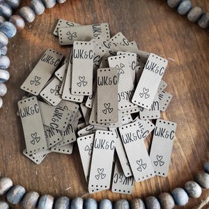 May include: A collection of rectangular beige faux leather tags featuring the text WK&C and three small heart outlines. Each tag has pre-punched holes at the top and bottom for sewing, arranged in a scattered pile on a wooden surface.