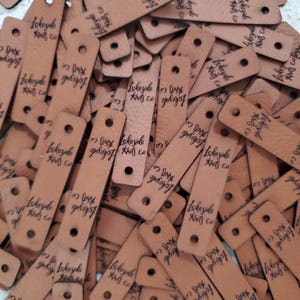 May include: A collection of rectangular tan leather tags, each featuring two pre-punched holes at the ends and the black stamped text Lakeside Knits Co. The tags are scattered in a pile, showing a smooth texture and consistent branding across all pieces.