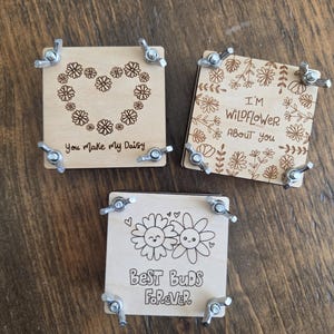 May include: Three square wooden flower presses with metal wing nuts at each corner. The top press features a daisy heart and the text "You make my Daisy". The second shows wildflowers and "I'M WILDFLOWER ABOUT YOU". The third shows two smiling flowers and "BEST BUDS FOREVER".