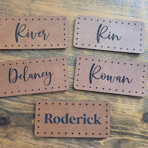 May include: Five rectangular tan leather labels with pre-punched holes along the edges. Each label features a black printed name in a cursive script font, displaying the names River, Rin, Delaney, Rowan, and Roderick on individual pieces.