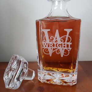 May include: A clear glass decanter with a square body and a stopper. The decanter is filled with a brown liquid and has a monogram etched on the front that reads "W WRIGHT".