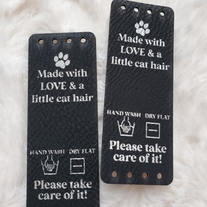 May include: Two rectangular black leather tags with silver text and a paw print icon. The text reads: Made with LOVE & a little cat hair, HAND WASH, DRY FLAT, and Please take care of it! Each tag features four pre-punched holes at the top and bottom.