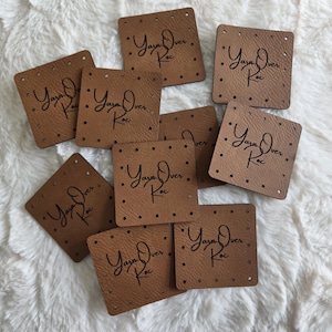 May include: Several square, brown leather tags with rounded corners and small holes around the edges. Each tag displays the text "Yaya Over Koc" in a handwritten font. The tags are arranged on a fluffy, white surface.