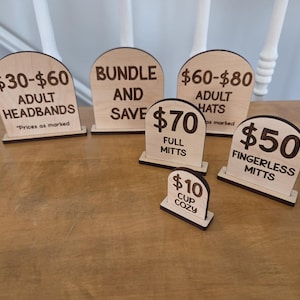 Peut inclure: Un ensemble de six panneaux en bois clair en forme d'arche avec du texte gravé au laser brun foncé. Les panneaux affichent divers prix et étiquettes de produits, notamment $30-$60 Adult Headbands, Bundle and Save, $60-$80 Adult Hats, $70 Full Mitts, $50 Fingerless Mitts et $10 Cup Cozy.
