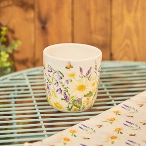 May include: A white ceramic mug decorated with a floral pattern of yellow daisies, purple lavender, and bees. The mug is cylindrical and rests on a light blue metal table. A matching floral cloth is also visible.
