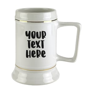 May include: White ceramic beer stein with a gold trim around the top and bottom. The stein has a handle and says "YOUR TEXT HERE" in black lettering.