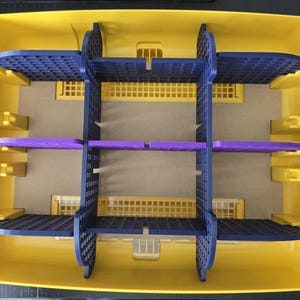 May include: A yellow plastic storage container with a grid of blue and purple dividers. The container has a rectangular shape with rounded corners and a flat base. The dividers create multiple compartments.