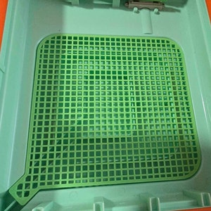 Puede incluir: Una rejilla de plástico verde con un patrón cuadrado, probablemente utilizada como bandeja o inserto para un pequeño juguete o proyecto artesanal.