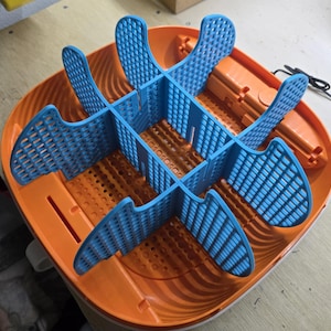Puede incluir: Un organizador de plástico naranja y azul con un diseño ranurado. El organizador tiene una base circular con una serie de divisores azules curvos que crean múltiples compartimentos. Los divisores están hechos de un material similar a una rejilla, y la base tiene una serie de pequeños agujeros.