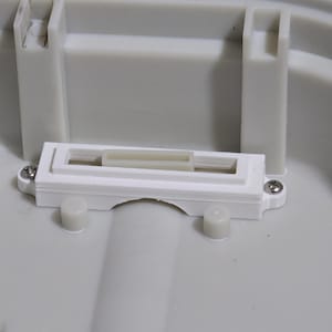 Puede incluir: Primer plano de un componente de plástico blanco con un marco rectangular y una pequeña pieza rectangular de color claro en el centro. El componente está fijado a una estructura de plástico gris claro más grande con dos tornillos.