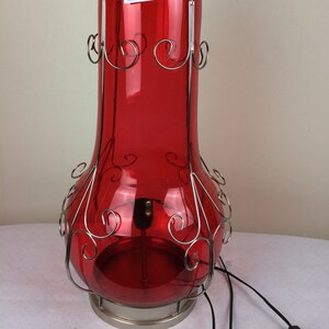 Peut inclure: Une lampe en verre rouge avec une cage en métal et un sommet argenté. La lampe a un design décoratif et est éclairée de l'intérieur.