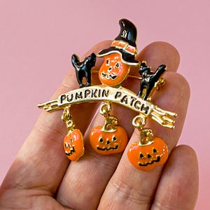 Könnte beinhalten: Eine Brosche aus goldfarbenem Metall mit einem Kürbisgesicht, das einen schwarzen Hexenhut trägt, flankiert von zwei schwarzen Katzen. Ein Banner trägt die Aufschrift PUMPKIN PATCH, mit drei hängenden Kürbis-Anhängern aus orangefarbenem Emaille.
