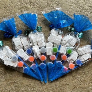May include: A set of six paint your own kits, each containing a small white ceramic truck, a paintbrush, and a small pot of paint. The kits are packaged in clear plastic bags with blue tulle and tied with a ribbon.