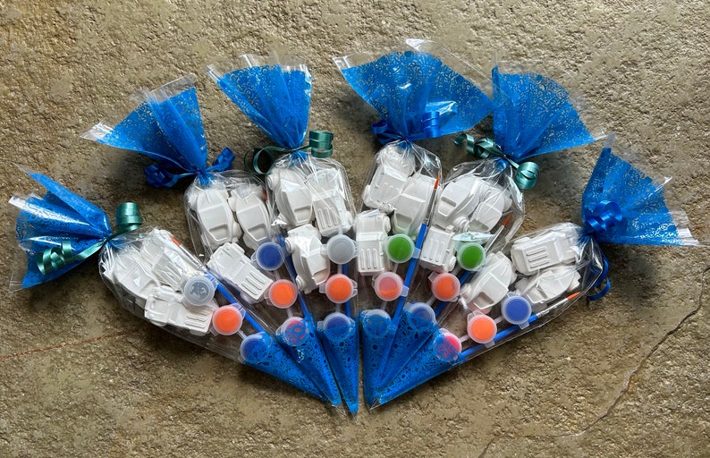 May include: A set of six paint your own kits, each containing a small white ceramic truck, a paintbrush, and a small pot of paint. The kits are packaged in clear plastic bags with blue tulle and tied with a ribbon.