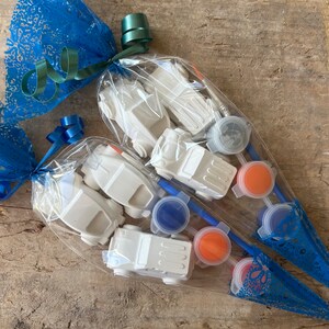 May include: Two clear plastic bags filled with white toy cars and paint pots. The bags are tied with blue and green ribbon and wrapped in blue lace.