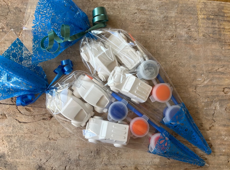 May include: Two clear plastic bags filled with white toy cars and paint pots. The bags are tied with blue and green ribbon and wrapped in blue lace.