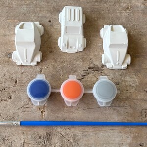 May include: A set of three white ceramic car figurines with three small paint pots and a paintbrush. The paint pots are blue, orange, and gray.