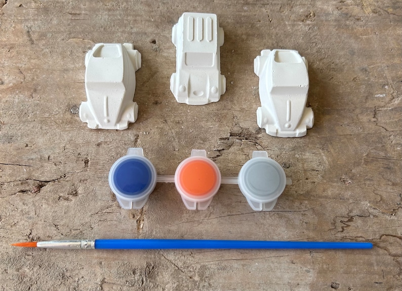 May include: A set of three white ceramic car figurines with three small paint pots and a paintbrush. The paint pots are blue, orange, and gray.