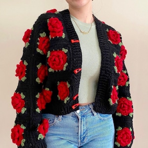 Op de afbeelding: Een zwart gebreid vest met rode en groene 3D-rozen. Het vest heeft een knoopsluiting en is getailleerd.
