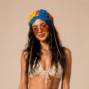May include: A beige crochet bikini top and matching wrap skirt with long fringe trim, paired with a blue, yellow, and orange floral crochet headpiece, brown leather cowboy boots, and a brown leather belt featuring large circular silver metal conchos.