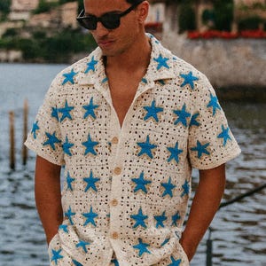May include: Cream-colored crochet shirt and shorts set with blue star patterns. The short-sleeved shirt has a button-down front and a collar. The shorts have pockets. The outfit is worn with sunglasses and sandals.