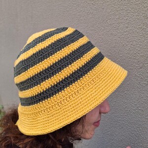 Op de afbeelding: Handgemaakte gehaakte bucket hat met afwisselende horizontale strepen in geel en grijs. De hoed heeft een brede rand en is gemaakt van zacht, getextureerd garen. De hoed is een stijlvol accessoire.