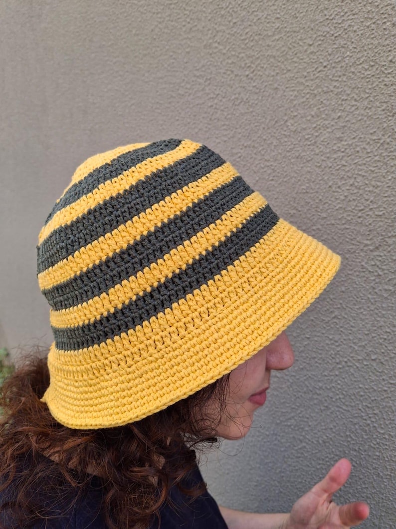 Op de afbeelding: Handgemaakte gehaakte bucket hat met afwisselende horizontale strepen in geel en grijs. De hoed heeft een brede rand en is gemaakt van zacht, getextureerd garen. De hoed is een stijlvol accessoire.
