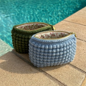 May include: Two handmade crochet pouches, one in olive green and the other in light blue, sit side-by-side. Each pouch features a textured, bobble stitch design, a floral patterned lining, and a gold zipper. The pouches are displayed outdoors.