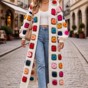 Puede incluir: Un cárdigan largo de crochet color crema con cuadrados florales coloridos. El cárdigan tiene una parte delantera abotonada y mangas largas. Las flores son en tonos de rojo, rosa, naranja, amarillo, azul y verde. La persona lleva vaqueros azules y una camiseta blanca.